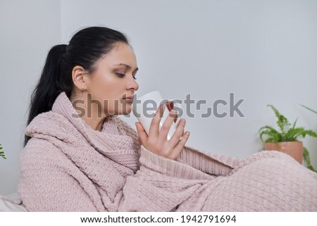 Beautiful woman sitting at home under knitted warm blanket with cup of tea
