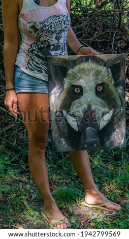 Outdoor natural environment with a female artist hodling her atrwotk piece. Acrilic canvas painting of a head of wild boar. 