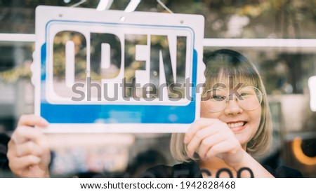 Young Asian woman hanging open sign by glass window. Female shop owner holding open notice in front of store