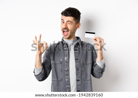 Very good. Smiling guy with plastic credit card showing okay sign, smiling and winking satisfied, recommend bank, standing on white background