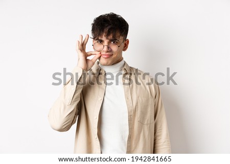 Smiling cunning guy in glasses shut his mouth on zipper, showing seal gesture near lips and smiling devious, keeping a secret, standing on white background