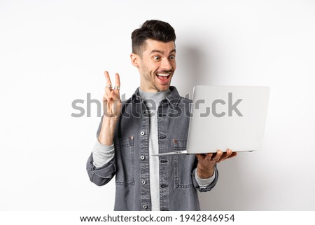 Funny young man video chat on laptop, showing v-sign and smiling at computer camera, standing against white background