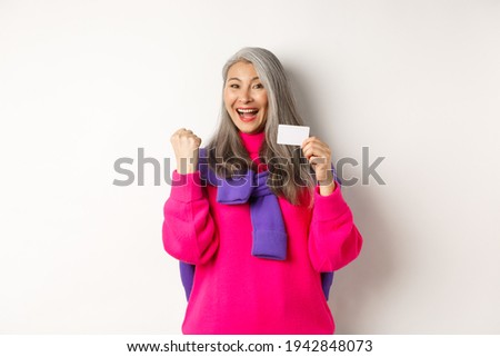 Shopping concept. Cheerful asian senior woman winning prize from bank, showing fist pump gesture and plastic credit card, standing happy over white background