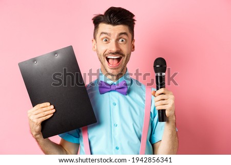 Holidays concept. Happy young man entertainer smiling at camera, holding clipboard and microphone, making speech on party event, standing over pink background