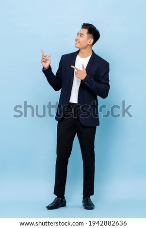 Full length portrait of happy smiling young handsome southeast Asian businessman pointing and looking upward on light blue studio background Royalty-Free Stock Photo #1942882636