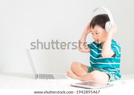 Asian boy student wear wireless headphone study online with video call teacher, Toddler boy child online learning listen english class.write notes look at laptop home.Covid19.Education.listen music.