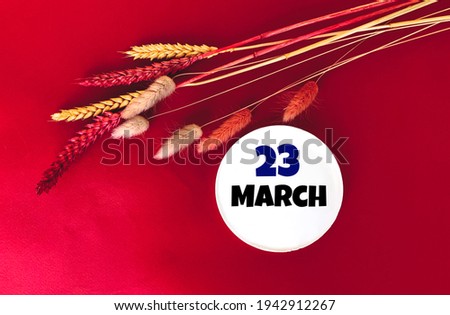 March 23 on a white round object .Next to it are sprigs of wheat on a red background.Calendar for March .