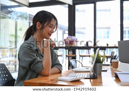 Side view of young woman designer sitting at creative workplace and looking at screen of tablet computer.