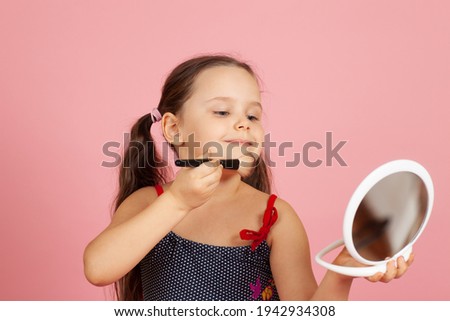 portrait of a smiling cute girl powdering her chin with a brush and looking at a mirror isolated on a pink background.