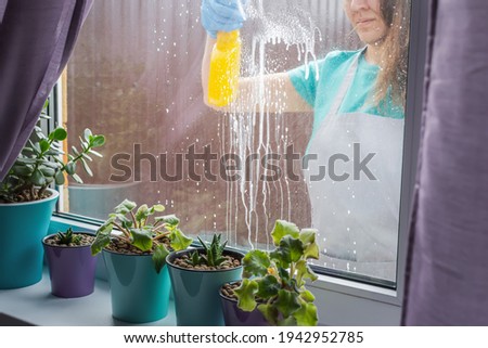 A woman in a work apron washes the windows of a house with a cleaning spray.Spring window washing. View through the glass from the inside.The concept of cleanliness in the house. Young woman housewife