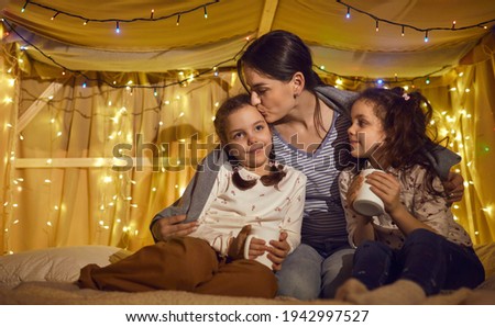Mother kissing with tenderness loving children daughters drinking hot tea or milk while spending time together in kids tent decorated with lights garland before bedtime. Cozy evening home atmosphere