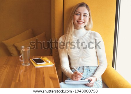 Beautiful young blonde student girl with long hair sitting in yellow interior cafe alone, studying, writing, painting in book, planner. Wearing white sweater and jeans, working with mobile phone. 