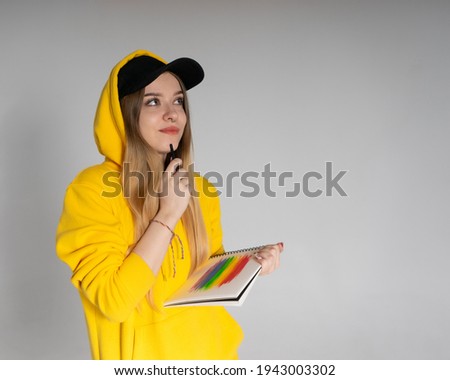 Happy woman with a smile in a yellow hoodie and black cap in a notebook draws a rainbow LGBTQ