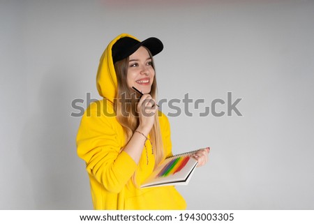 Happy woman with a radiant smile in a yellow hoodie and black cap in a notebook draws a rainbow LGBTQ