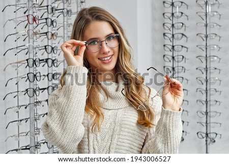 Woman chooses glasses in the store. Blonde in a beige sweater buys glasses. Girl on a background of shop windows with different models of glasses. Royalty-Free Stock Photo #1943006527
