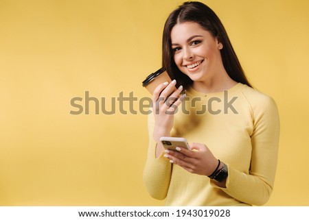 Pretty girl using phone and drink coffee on yellow background. Studio photoshoot of brunette female in yellow sweater. Happy smiled woman. Place for text