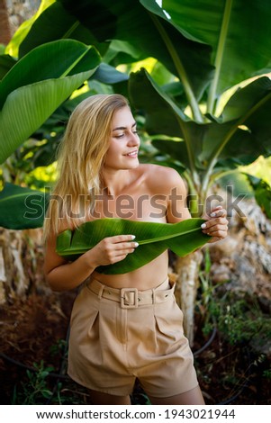 A beautiful young woman stands in a beautiful pose near a banana tree, her little girl covers her breasts with a leaf on a banana plantation.