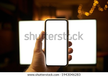 Mockup of hand holding modern smartphone with pc monitor on the background. Blank screen design template