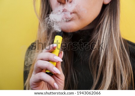 A young girl smokes disposable electronic cigarette. Bright yellow background Royalty-Free Stock Photo #1943062066