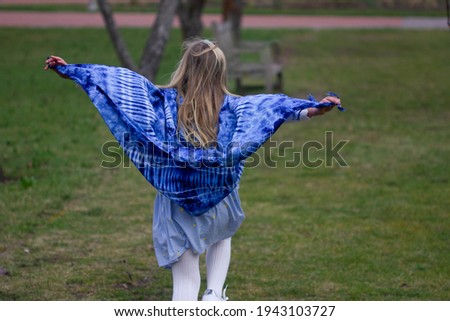 A girl is running with a bright blue scarf in a garden or city park. A blond model from the back, with her hands up on green grass background. Stock photography.