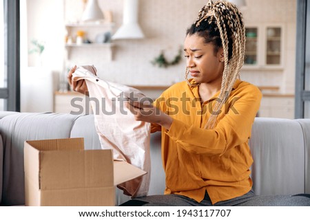 A confused sad african american girl unpacking her parcel at home. Surprised young woman got an unexpected order, girl is upset with her parcel, unsuccessful online shopping. Home delivery Royalty-Free Stock Photo #1943117707