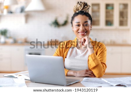 Satisfied good looking young African American stylish woman, freelancer, student or real estate agent, sitting at her desk at home office, looking at the camera and smiling pleasantly Royalty-Free Stock Photo #1943120119
