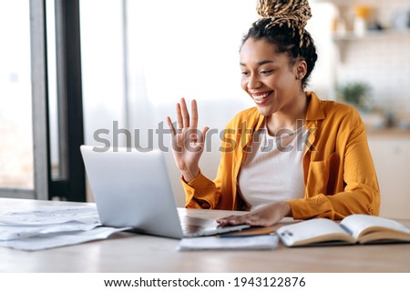 Online video communication. Joyful attractive African American woman, freelancer or student, in casual clothes, uses laptop for online video conference with colleagues or friends, greets, smiles Royalty-Free Stock Photo #1943122876