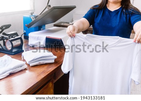 unrecognizable woman, holding white shirts behind a sublimation or screen printing machine for t-shirts. graphic design concept. Royalty-Free Stock Photo #1943133181