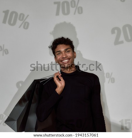 Smiling young african american guy looking at camera while standing in front of screen with percents on it and holding paper bags. Discount and sale concept