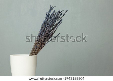 A bouquet of dry blue lavender stands in a white vase on a table against a gray wall. Space for text.