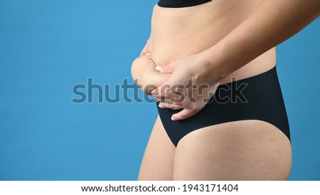 Cropped view of woman in black panties holds her belly fat on blue background. Body control, diet and weight loss concept. High quality photo