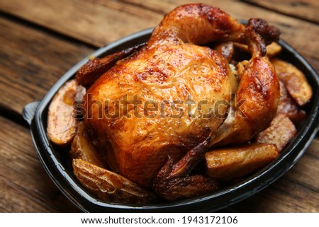 Delicious grilled whole chicken with potato in plastic container on wooden table, closeup. Food delivery service