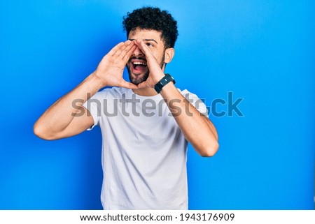 Young arab man with beard wearing casual white t shirt shouting angry out loud with hands over mouth 