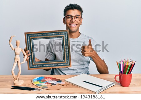 Young handsome african american man painter sitting at art studio holding empty frame smiling happy and positive, thumb up doing excellent and approval sign 