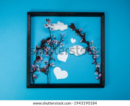Creative spring composition made of cherry blossom tree branches and wooden picture frame on blue background with heart shape and clouds, spring concept, picture in picture, flat lay, top view
