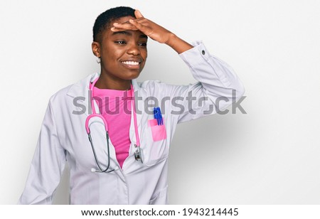 Young african american woman wearing doctor uniform and stethoscope very happy and smiling looking far away with hand over head. searching concept. 