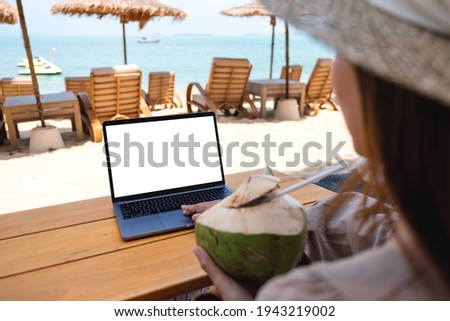 Mockup image of a woman using and touching on laptop touchpad with blank desktop screen while drinking coconut juice on the beach