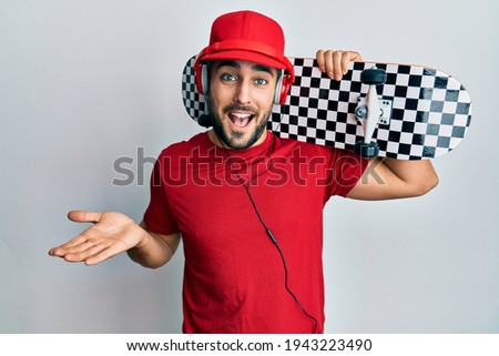 Young hispanic man using headphones holding skate celebrating achievement with happy smile and winner expression with raised hand 
