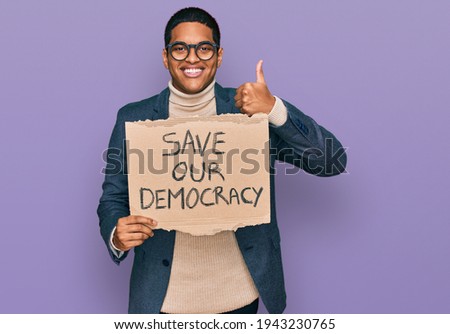 Young handsome hispanic man holding save our democracy protest banner smiling happy and positive, thumb up doing excellent and approval sign 