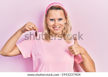 Beautiful caucasian blonde woman holding cockroach smiling happy and positive, thumb up doing excellent and approval sign 
