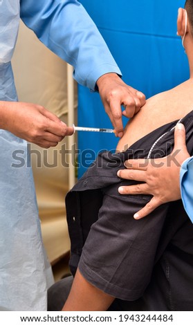 A female nurse is injecting vaccine to left shoulder of a young man. A close-up and selective focus photo of nurse hand, needle and syringe. A medical concept.