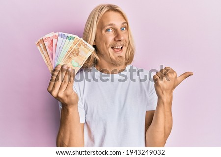 Caucasian young man with long hair holding philippine peso banknotes pointing thumb up to the side smiling happy with open mouth 
