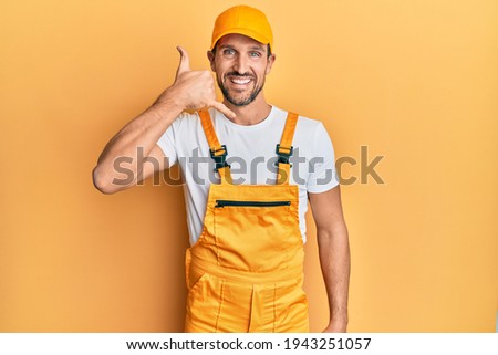 Young handsome man wearing handyman uniform over yellow background smiling doing phone gesture with hand and fingers like talking on the telephone. communicating concepts. 