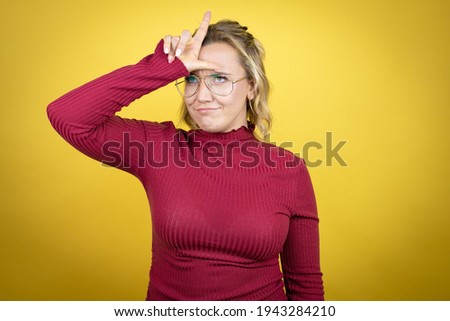 Young caucasian woman wearing casual red t-shirt over yellow background making fun of people with fingers on forehead doing loser gesture mocking and insulting.