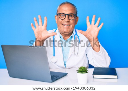Senior handsome man with gray hair wearing doctor uniform working using computer laptop showing and pointing up with fingers number ten while smiling confident and happy. 
