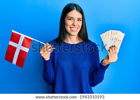 Young hispanic woman holding denmark flag and krone banknotes smiling with a happy and cool smile on face. showing teeth. 