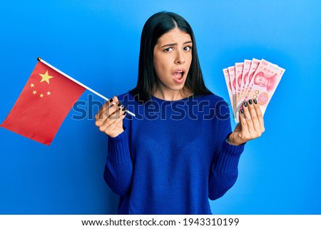 Young hispanic woman holding china flag and yuan banknotes clueless and confused expression. doubt concept. 