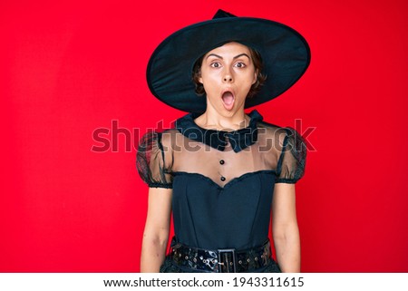 Young hispanic woman wearing witch halloween costume scared and amazed with open mouth for surprise, disbelief face 