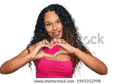 Young hispanic woman with curly hair wearing pink top smiling in love showing heart symbol and shape with hands. romantic concept. 