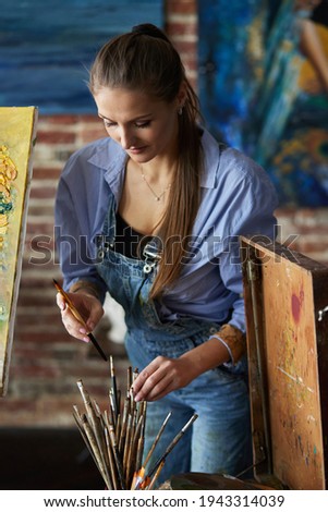 Painter choosing art brushes for work. Portrait of young smiling female artist in art studio. Creative process, relaxation, leisure, hobby, stress management.